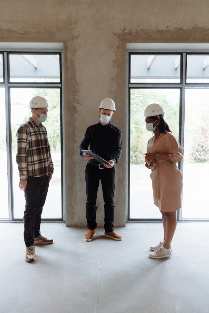 Three people in masks and hard hats discuss real estate indoors with natural lighting.
