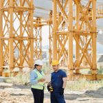 Two construction workers examining plans at a site with large steel structures.
